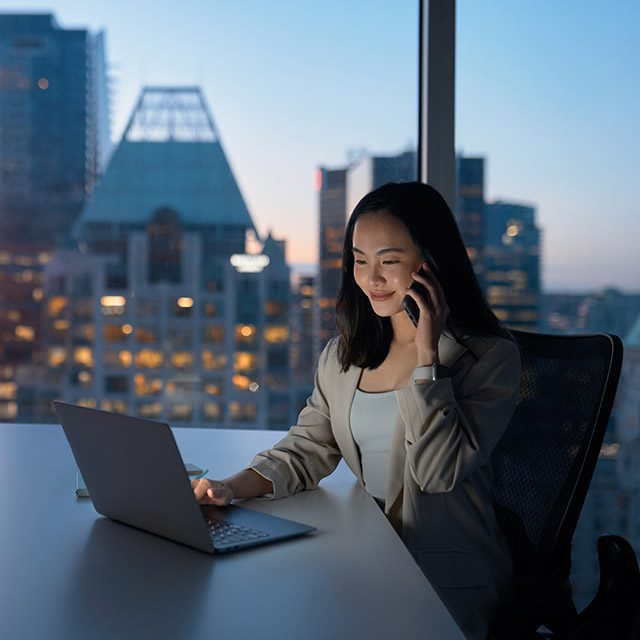 Young busy Asian business woman executive working on laptop making call at night in dark corporate office. Professional businesswoman manager talking to client using computer, city evening window view