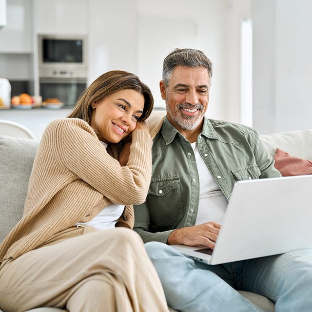 Happy middle aged couple using laptop relaxing on couch at home. Smiling mature man and woman looking at computer watching video, browsing or shopping in ecommerce store sitting on sofa in living room